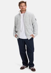 Grau gestrickte Zip-Jacke mit hohem Kragen und strukturiertem Muster. Trägt man über ein weißes Hemd, kombiniert mit marineblauen Hosen und dunklen Schuhen.