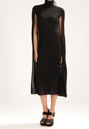 Kobieta ubrana w czarną midi sukienkę z wysokim kołnierzem i rękawami w stylu peleryny oraz czarne platformy na obcasie z paskami wokół kostek.