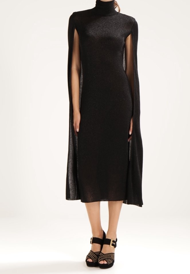 Femme portant une robe midi noire à col montant avec des manches style cape et des talons compensés noirs avec brides à la cheville.