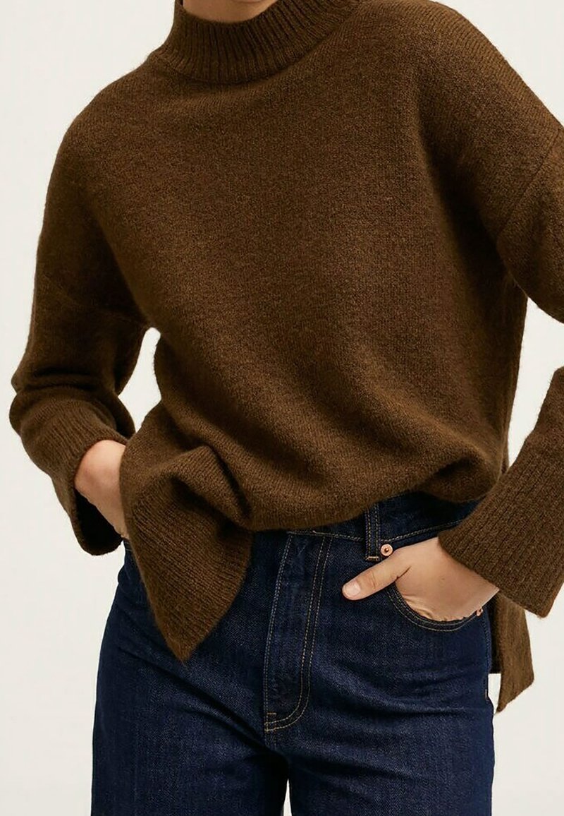 Personne portant un pull à col roulé côtelé marron à manches longues et un jean taille haute bleu foncé, les mains dans les poches.
