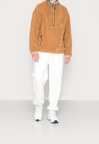 Persona che indossa un pullover di pile teddy marrone, pantaloni della tuta bianchi e sneaker Adidas bianche, in piedi contro uno sfondo chiaro e semplice.