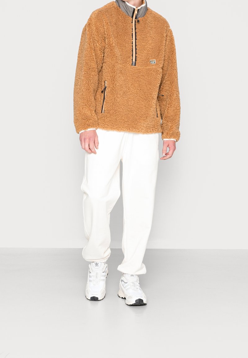 Persona che indossa un pullover di pile teddy marrone, pantaloni della tuta bianchi e sneaker Adidas bianche, in piedi contro uno sfondo chiaro e semplice.