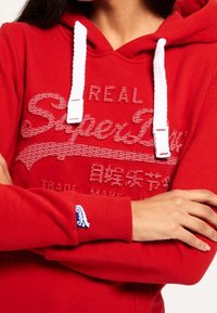 Rode hoodie met een gestructureerd logo in wit en lichtgrijs, voorzien van een kangoeroezak, trekkoordcapuchon en een blauw merkteken op de mouw.