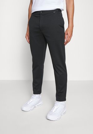 Mand iført sorte slim-fit bukser, hvide sneakers og en hvid skjorte, stående foran en ensartet lys grå baggrund.