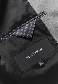 Juodos striukės pamušalas, kuriame matyti nedidelis raštuotas tamsiai mėlynas kišeninis rankšluostis, įdėtas viduje, ir etiketė "Martinique" su dydžiu EUR 50, JAV 40.