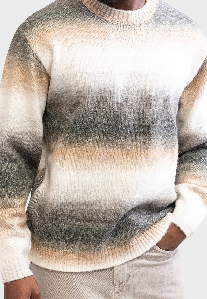 Homme portant un pull en maille dégradé beige, gris et blanc, avec la main dans la poche d'un pantalon beige, debout devant un fond clair.