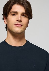 Marinblå crew-neck sweatshirt med en texturerad tyg. Har en liten logotyp på bröstet. Närbild av ett neutralt uttryck.