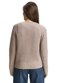 Woman with long brown hair wearing a beige ribbed knit sweater and blue jeans, viewed from the back against a white background.