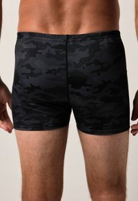 Homme portant un short de bain noir et gris foncé avec un motif camouflage, vu de l'arrière bas jusqu'à mi-cuisse.