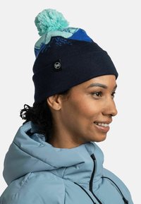 Gorro de punto con un pompón turquesa, patrón geométrico de montañas en azul y marino, con una etiqueta negra en el costado.