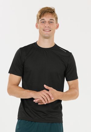 Jeune homme aux cheveux blonds courts souriant, portant un maillot de sport noir à manches courtes et un short bleu sarcelle, debout les mains jointes.