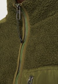 Närbild av en olivgrön fleecejacka med texturerad tyg och dragkedja fram med en svart- och grön dragrem.