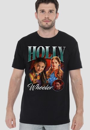 Mann trägt ein schwarzes T-Shirt mit drei Porträts eines Mädchens und dem Text "Holly Wheeler" vor einem einfarbigen grauen Hintergrund.