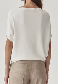 Blusa blanca de corte holgado y mangas cortas, combinada con pantalones beiges a rayas. Textura de tela suave, detalles mínimos, colores neutros.