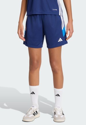Persona con pantalones cortos deportivos Adidas azul marino, calcetines blancos Adidas hasta el tobillo y zapatillas blancas con rayas negras, de pie con las manos relajadas.