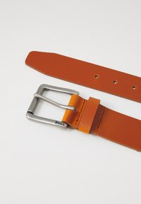Ceinture en cuir orange avec une boucle en métal argenté. Comprend deux sections : une large extrémité et une queue étroite avec des trous pour l'ajustement. Le logo de la marque est présent.