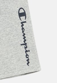 Γκρι κοντομάνικο πουκάμισο από απαλό ύφασμα, με εμφανές λογότυπο "Champion" σε μπλε ναυτικό χρώμα κατά μήκος της πλευράς, γραμμένο με χειρόγραφο στυλ.