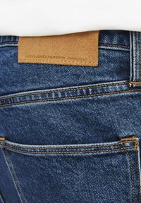Jeans din denim albastru cu o etichetă din piele maro pe care scrie "JACK & JONES" la talie. Prezintă cusături vizibile și două buzunare la spate.