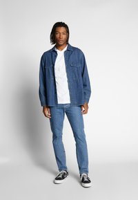 Denim-Overshirt in Mittelblau mit zwei Brusttaschen, über einem weißen T-Shirt getragen, kombiniert mit hellblauen Jeans und schwarzen Sneakers.
