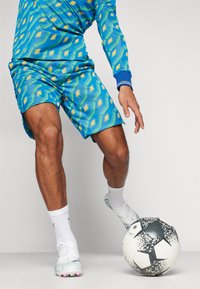 Blau- und grün gemustertes Sportoberteil und Shorts, kombiniert mit weißen Fußballschuhen und Socken, neben einem schwarz-weißen Fußball.