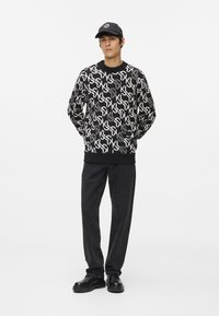 Melnā un baltā raksturā adīta džemperis ar grafisku dizainu, apvienots ar melnajām biksēm un melnām kurpēm. Melnā cepure noslēdz apģērbu.