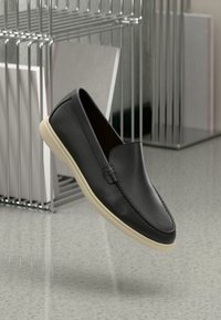 Mocassin en cuir noir avec semelle beige, présenté flottant au-dessus d'un sol moucheté avec une étagère métallique et des livres en arrière-plan.