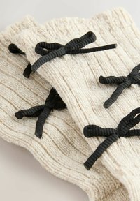 Knit wrist warmers in cream with a ribbed texture, featuring black ribbon ties for closure, shown stacked against a neutral background.