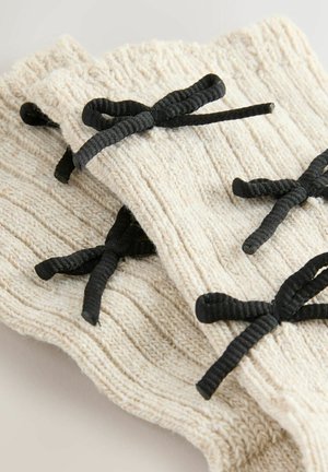 Strick-Handgelenkwärmer in creme mit gerippter Textur, die mit schwarzen Bändern zum Binden geschlossen werden. Sie sind übereinander gestapelt vor einem neutralen Hintergrund zu sehen.