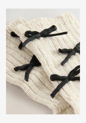 Kreemivärvi soonikud randme soojendajad, ribakujulise tekstuuriga, mustade paeltega sulgemiseks, asetatud neutraalsetes toonides taustale.