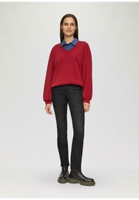 Vrouw die staat, gekleed in een rode V-halstrui over een blauwe overhemd, zwarte jeans en zwarte chunky laarzen, tegen een effen achtergrond.