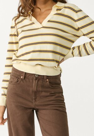 Woman wearing cream sweater with brown and yellow horizontal stripes and high-waisted brown pants, hand on hip against plain background.