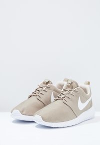 Ett par beige Nike löparskor med vita sulor och vita swoosh-loggor, uppställda på en vit yta mot en ljus bakgrund.