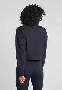 Vrouw met krullend haar, met de rug naar de kijker, gekleed in een zwarte cropped sweatshirt en donkere strakke leggings tegen een eenvoudige lichte achtergrond.