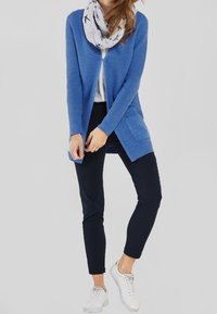 Blauwe geribde cardigan met open voorkant en zakken, gedragen met een grijs shirt, marineblauwe broek en witte sneakers. Sjaal heeft een vogelpatroon.