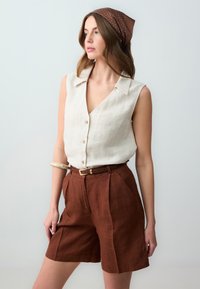 Camicetta senza maniche beige chiaro con colletto e bottoni, abbinata a pantaloni corti in lino marrone con pieghe e una cintura color sabbia. Decorata con un foulard a fantasia.
