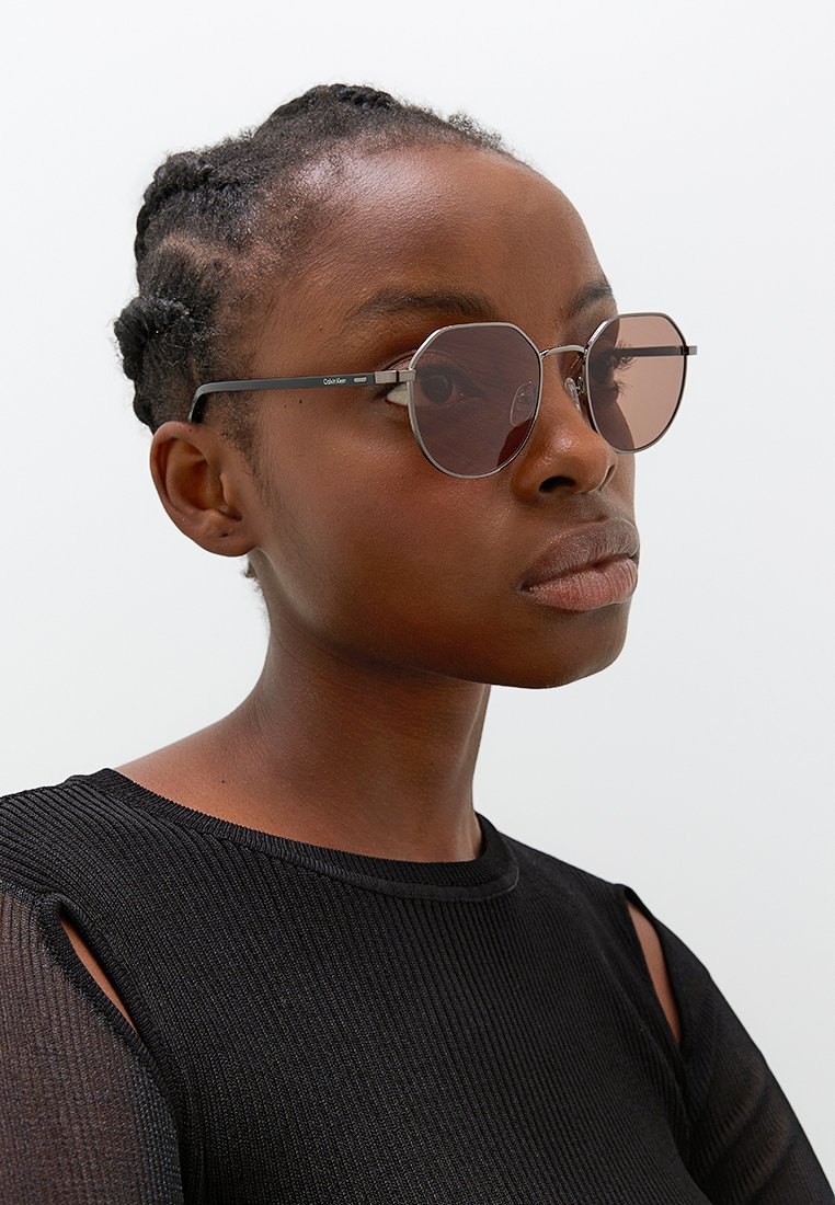 Occhiali da sole rotondi di colore rosa con una sottile montatura in argento. Le lenti sono di grande dimensione, con un design minimalista, poggiate su una superficie superiore scura e texturizzata.