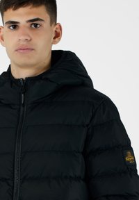 Giacca a vento nera con cappuccio, caratterizzata da sezioni trapuntate, una zip frontale e una toppa con logo sulla manica sinistra. Texture liscia e leggera.