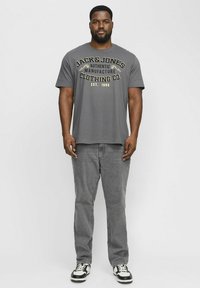 Graues T-Shirt mit gelbem und schwarzem grafischen Text, kombiniert mit grauen Jeans und schwarz-weißen Sneakers. Lässiger Schnitt, kurze Ärmel.