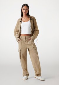 Beige gekreukt stoffen jack met een trekkoord en bijpassende cargobroek, voorzien van zijzakken, gecombineerd met een witte cropped tanktop en witte sneakers.