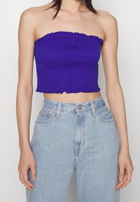 Lila off-shoulder crop top med smockad textur och volangkant, kombinerad med högmidjade ljusblå jeans med koppardetaljer.