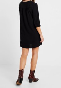 Robe noire à manches courtes avec une coupe ample et un détail froncé à l'arrière, associée à des bottines marron en cuir verni à talon carré.