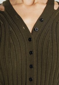 Olivgrön stickad topp med vertikal ribbning, featuring V-ringning och tre svarta knappar. Utmärkande utskurna axeldetaljer.