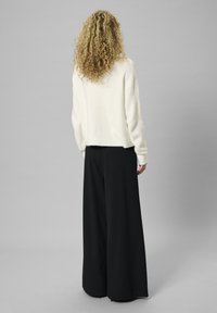 Häkelstrukturierter, cremefarbener Pullover mit lockerem Schnitt und geripptem Saum, kombiniert mit weit geschnittenen, schwarzen Hosen.