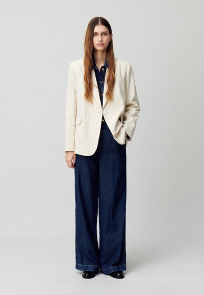 Blazer a righe crema con chiusura a un bottone, abbinato a una camicia di jeans scuri e pantaloni larghi in denim scuro; scarpe nere completano il look.