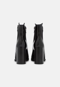 Bottines noires avec une finition brillante, dotées d'un talon épais et d'une fermeture éclair à l'arrière, au design épuré sans motifs visibles.