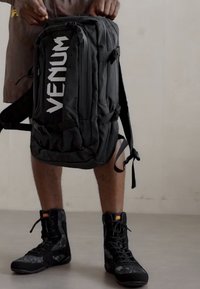 Sac à dos noir Venum avec un grand logo blanc, plusieurs compartiments, tissu texturé, bretelles réglables et conception durable, tenu par une personne portant des bottes.