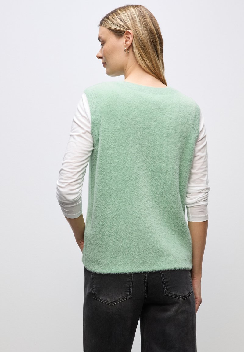 Miętowy, puszysty, bezrękawnik nałożony na biały, długi rękaw, w połączeniu z ciemnoszarymi spodniami. Gładka tekstura, okrągły dekolt i luźny krój.