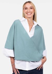 Uśmiechnięta kobieta ubrana w luźny, jasnoniebieski sweter na zakładkę noszony na białej koszuli z podwiniętymi rękawami, pozująca z rękami w kieszeniach.