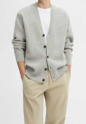 Mand iført lys grå ribbet cardigan med knapper over hvid t-shirt og beige bukser, hænderne i lommerne, mod en ensfarvet baggrund.