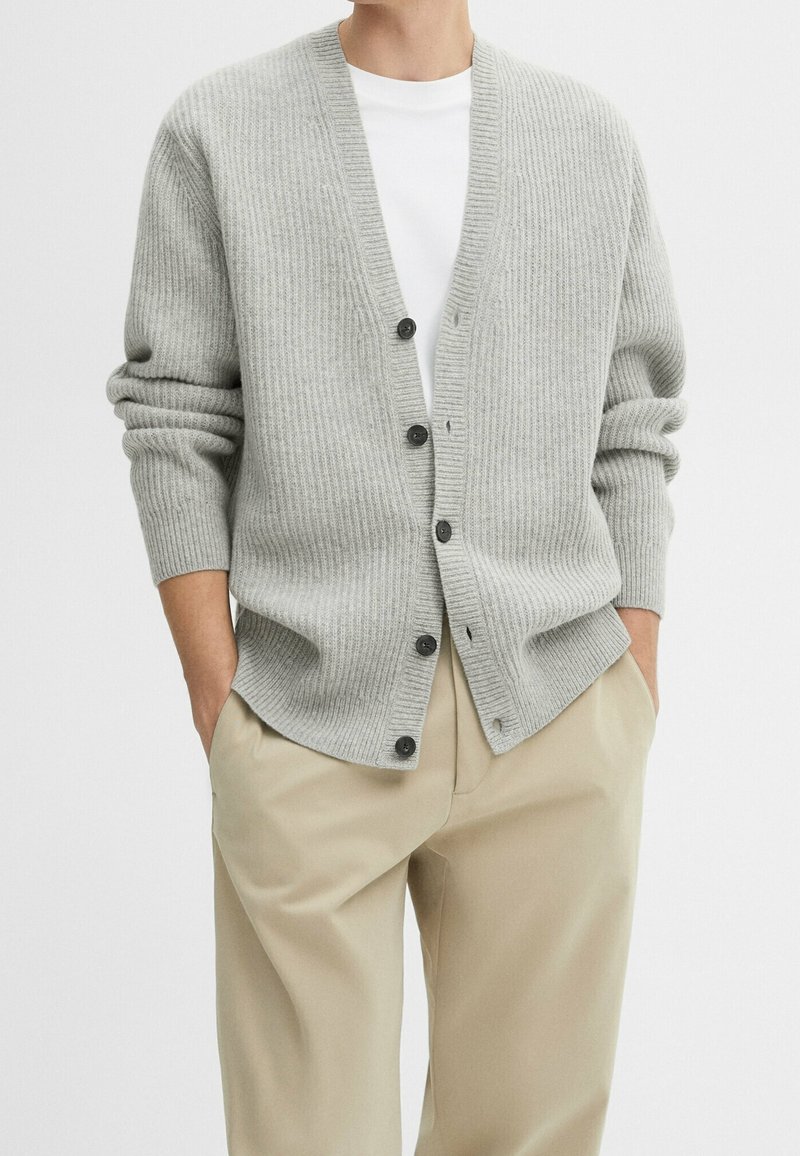 Homme portant un cardigan gris clair à côtes boutonné sur un t-shirt blanc avec un pantalon beige, les mains dans les poches, sur un fond uni.